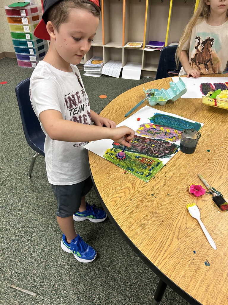 A boy painting at a table in a classroom. A girl sits behind him. He wears a hat and sneakers.