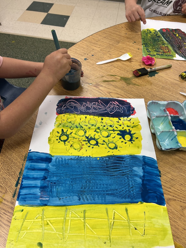 A person uses a paintbrush to apply paint to a large canvas with blue, yellow, and green colors.