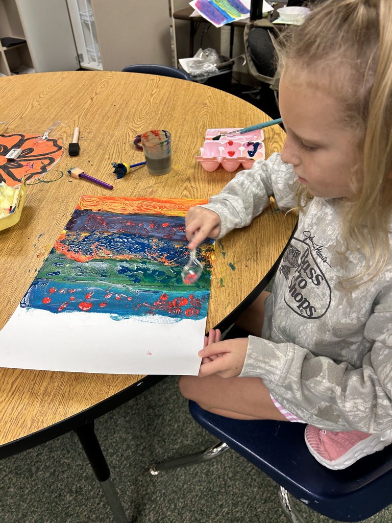 A child with blonde hair, seated at a round wooden table, is engaged in painting with multicolored paints.