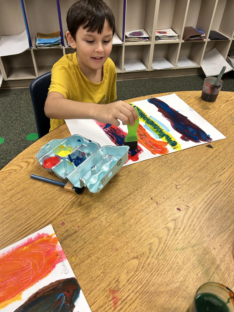 A young boy sitting at a table, painting with a brush. He has a palette and a cup.