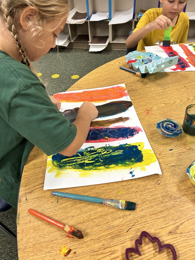 Two children are painting on a wooden table, one wearing green and the other yellow.