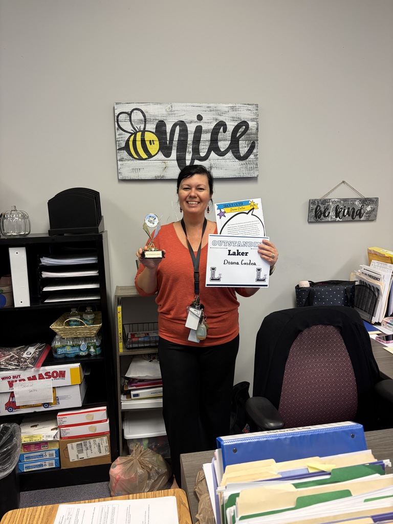 A woman in an orange shirt holds a certificate and an award in an office with shelves and a chair.