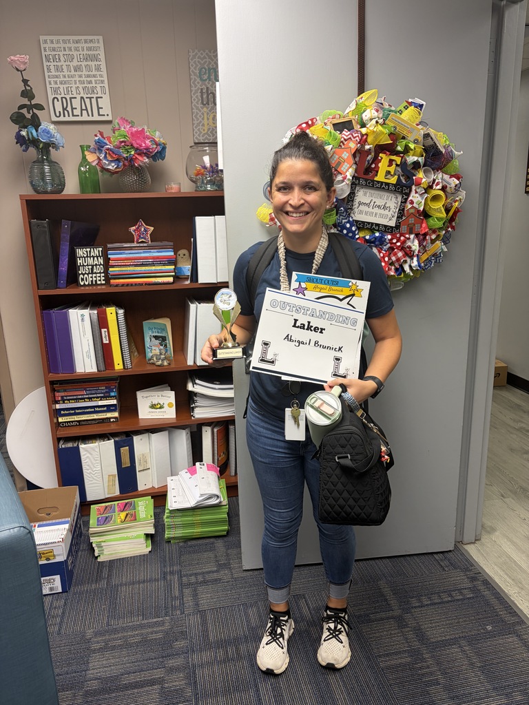 A woman stands with a certificate and trophy in a room with a bookshelf and decorative wreath.