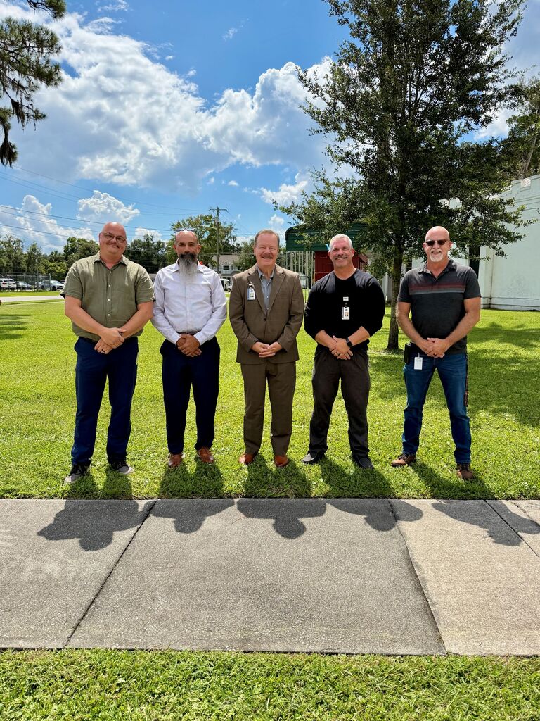 Five men stand in a line on a sidewalk, dressed in various colored shirts and pants. Behind them is a grassy area with a tree and a building.