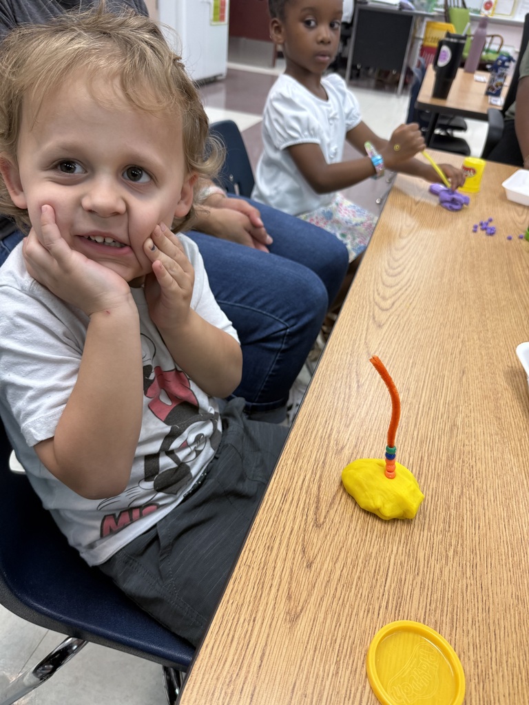 A young boy smiles at the camera. In front of him, there is a table with a creation made of yellow dough and beads.