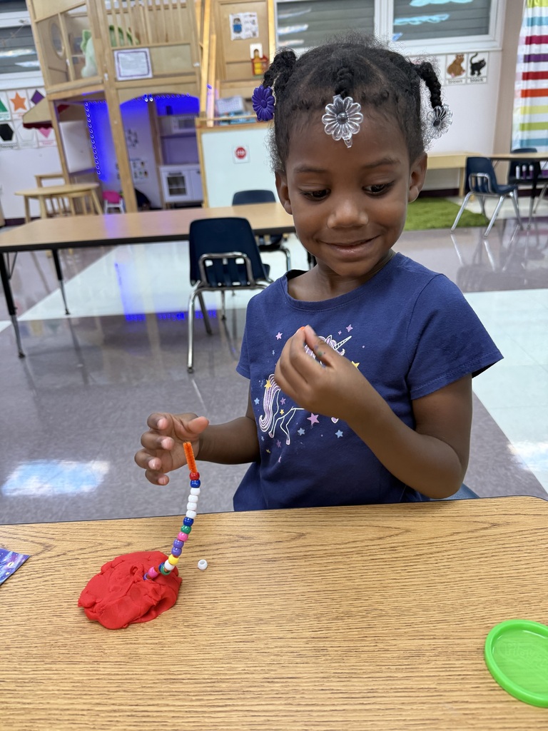 A child sits at a table in a classroom, engaged in an activity with colorful beads.