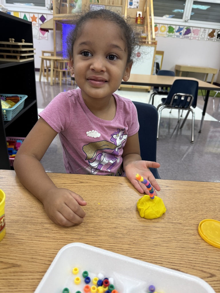 A young girl sitting at a table creating with yellow dough and beads.