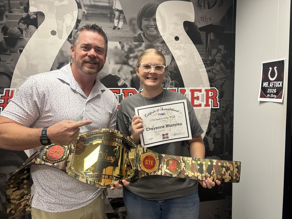 Two people pose with a large gold championship belt and a certificate against a horseshoe backdrop.
