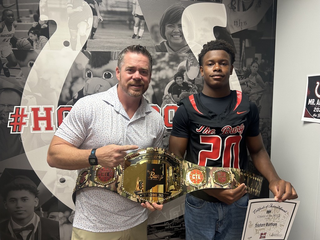 Two men in front of a large sports poster. One man holds a championship belt, the other holds a certificate.