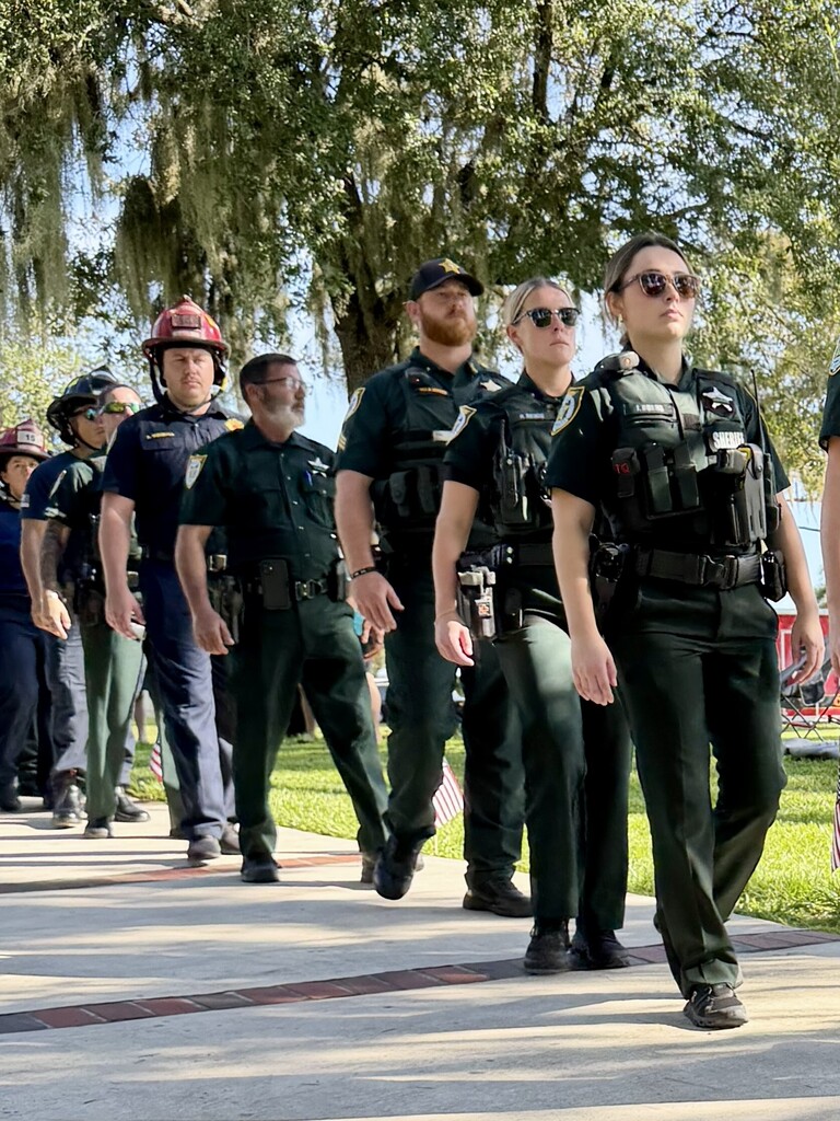 Six uniformed officers, three in helmets, walk on a sidewalk. Behind them, trees and grass.