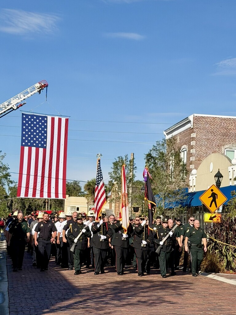 A line of uniformed individuals marches outdoors with flags. American flag hangs above, buildings and greenery on either side.