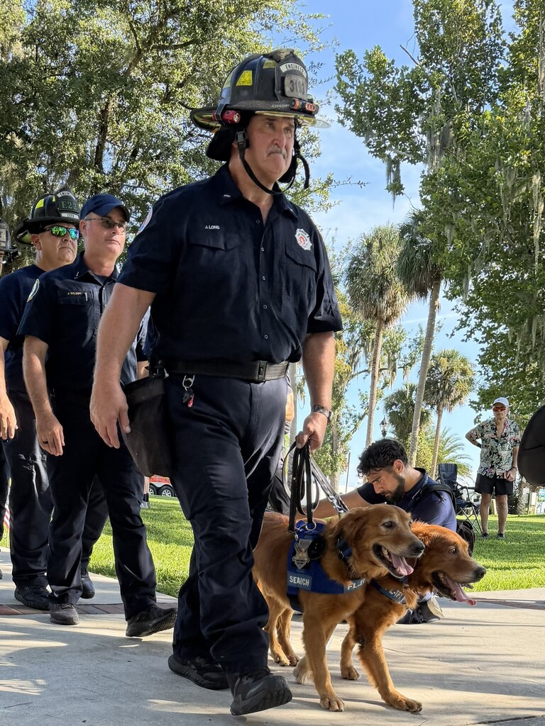 A firefighter, wearing a helmet and holding a leash, walks two dogs on a sidewalk. Other firefighters stand behind.