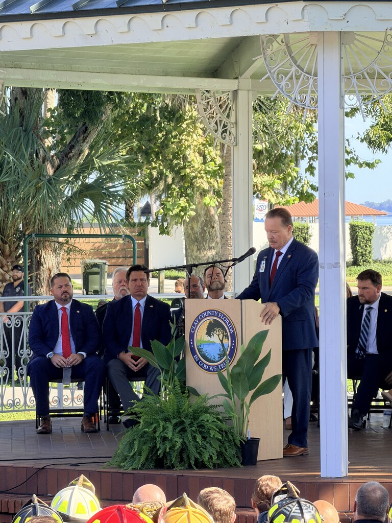 A man speaks at a podium in a gazebo. Men in suits sit behind him. Trees and a building are in the background.