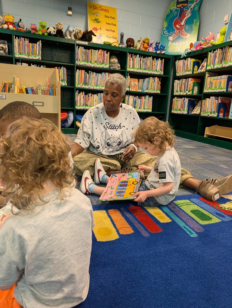 Woman sits on floor in front of children, reading a book. Colorful carpet with musical notes. Library in background with shelves full of books.