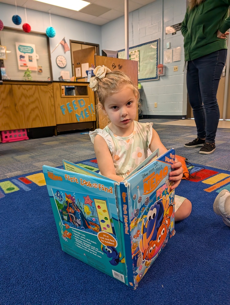 A young child sits on a floor with a blue rug, reading a colorful book. A person stands behind them.
