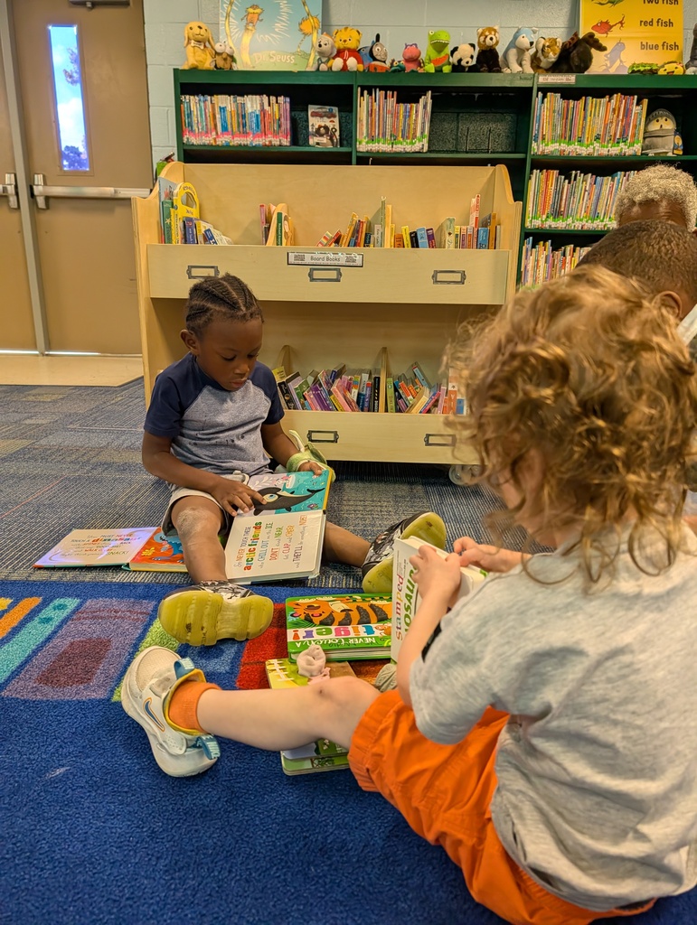 Two children sit on a blue carpeted floor, sharing books. Behind them are shelves filled with books and toys.