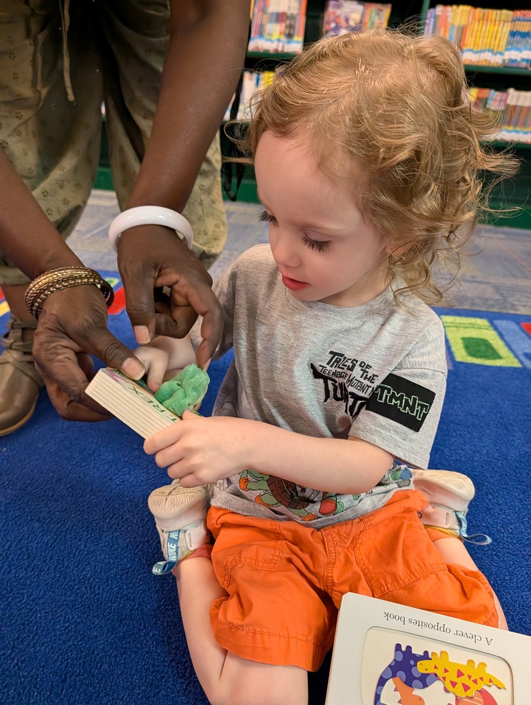 A child in an orange shirt is holding a book while another adult helps them. Background shows bookshelves.