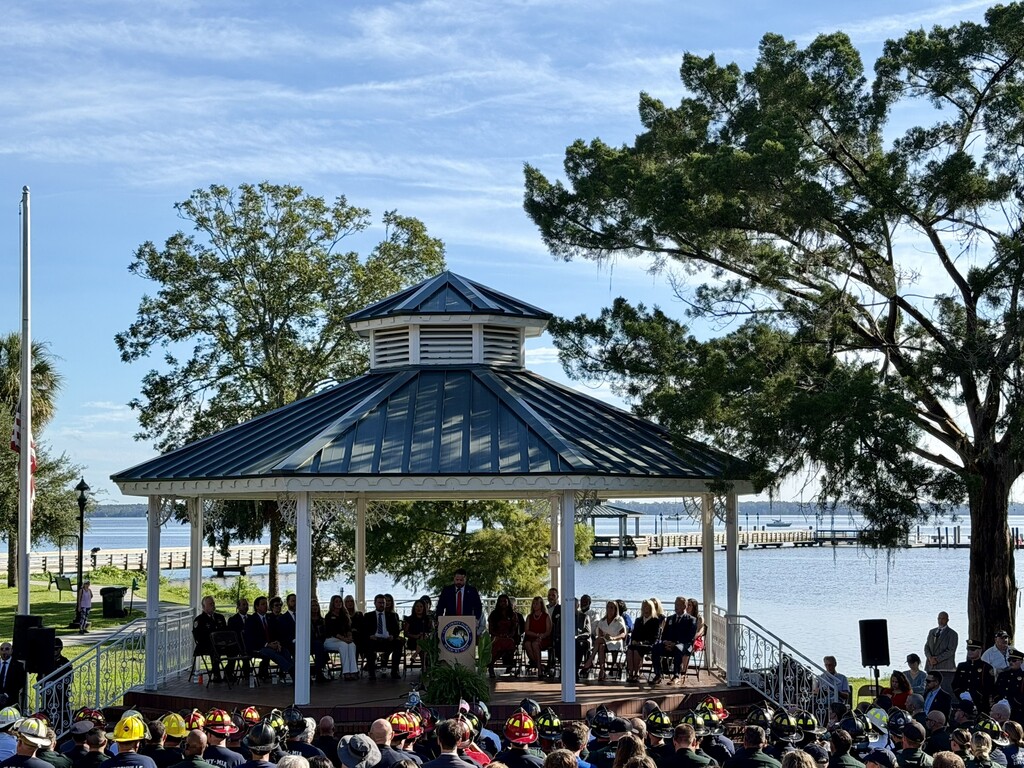 A crowd of people are seated at tables under a gazebo with a lake in the background.