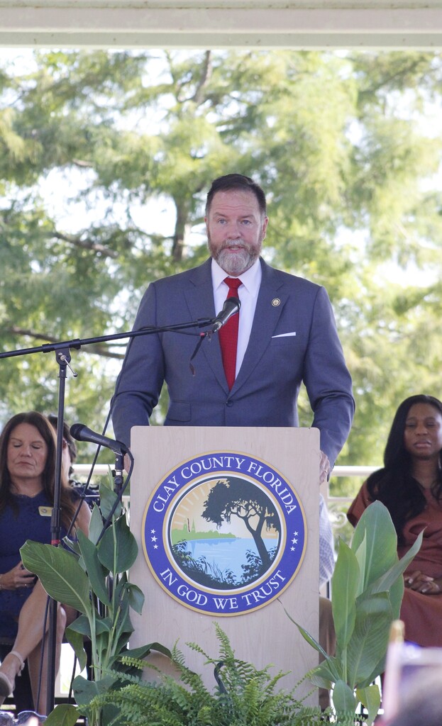 A man in a suit and tie at a podium with a microphone and Clay County Florida logo.