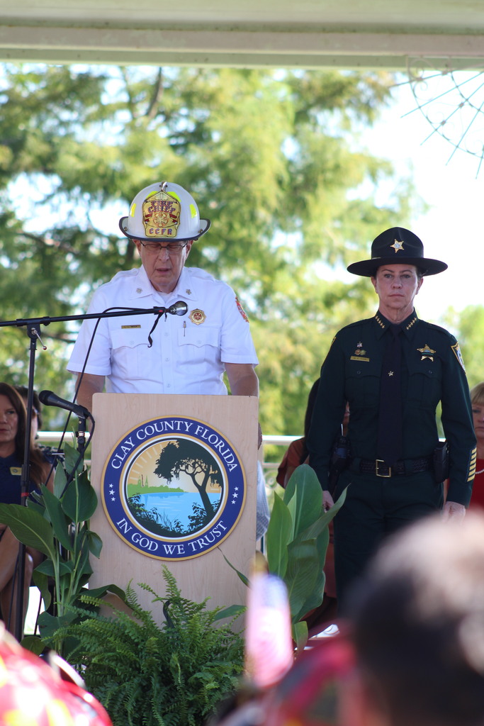 A person stands at a podium with a logo behind them. A uniformed individual stands nearby. A crowd sits in the background.