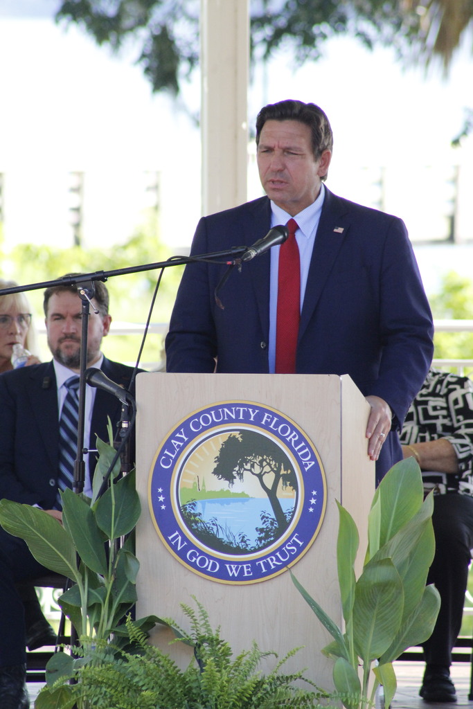A man speaking at a podium. Behind him, an emblem on the podium reads "Clay County Florida."