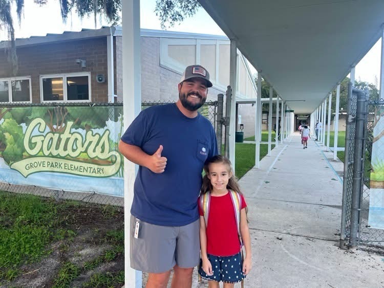 Man and child standing in front of an elementary school building, man giving a thumbs-up.