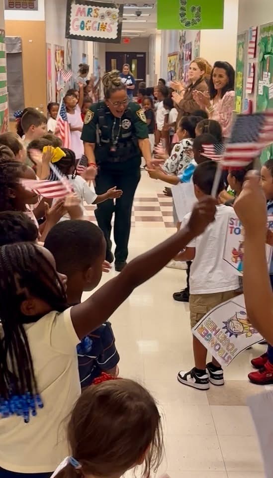 Children waving flags, one holding a sign. A police officer walks through a school hallway. Bright hallway with decorations.