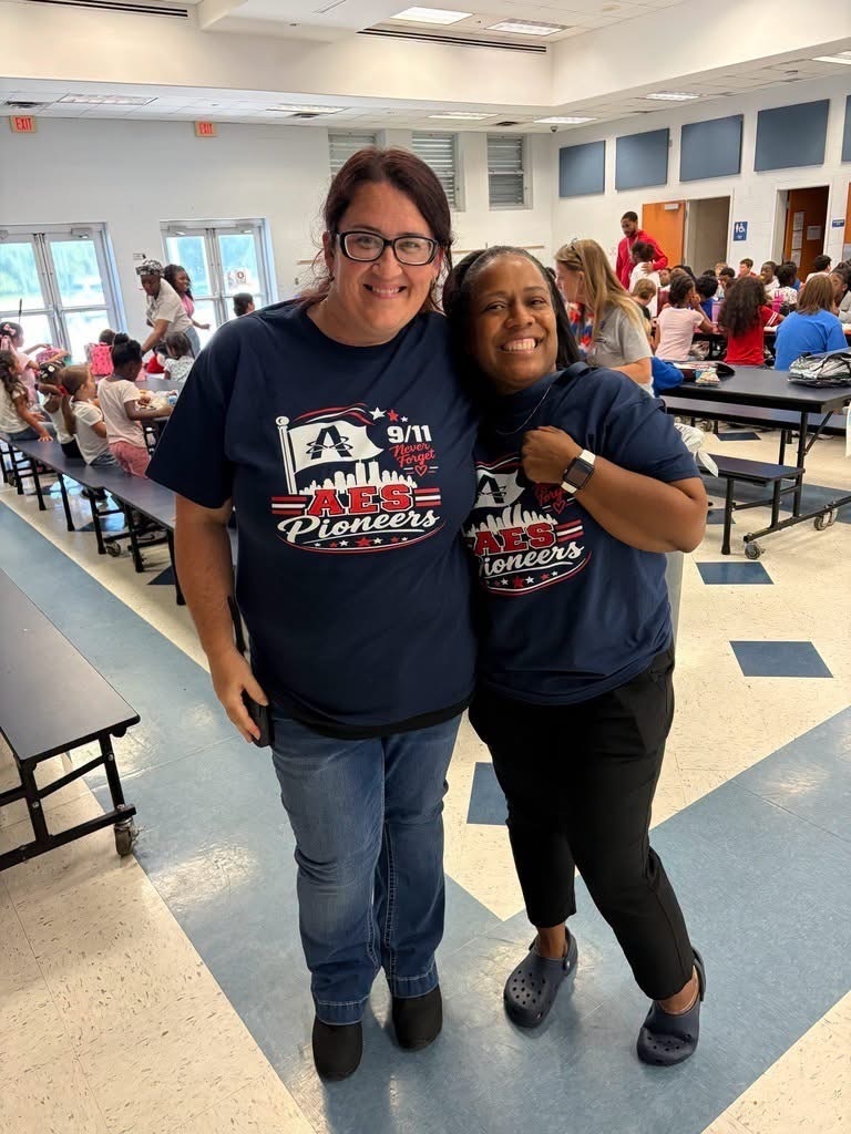 Two women stand in a room with blue floors and tables, smiling for a photo. Their shirts say "AES Pioneers."