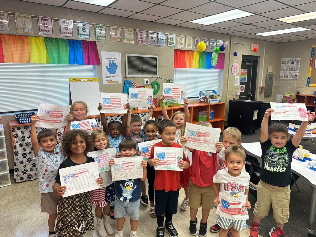A classroom with a colorful background; young children hold up small paper flags.