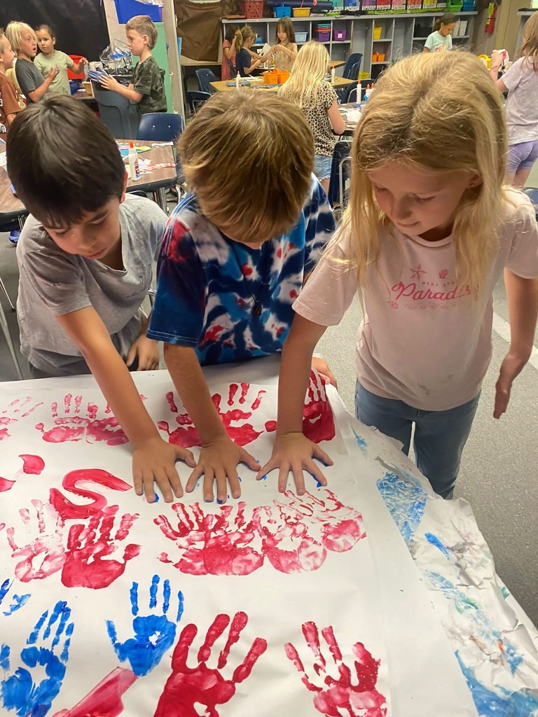 Three children work together on a table covered with blue and red handprints. Behind them, people work at tables.