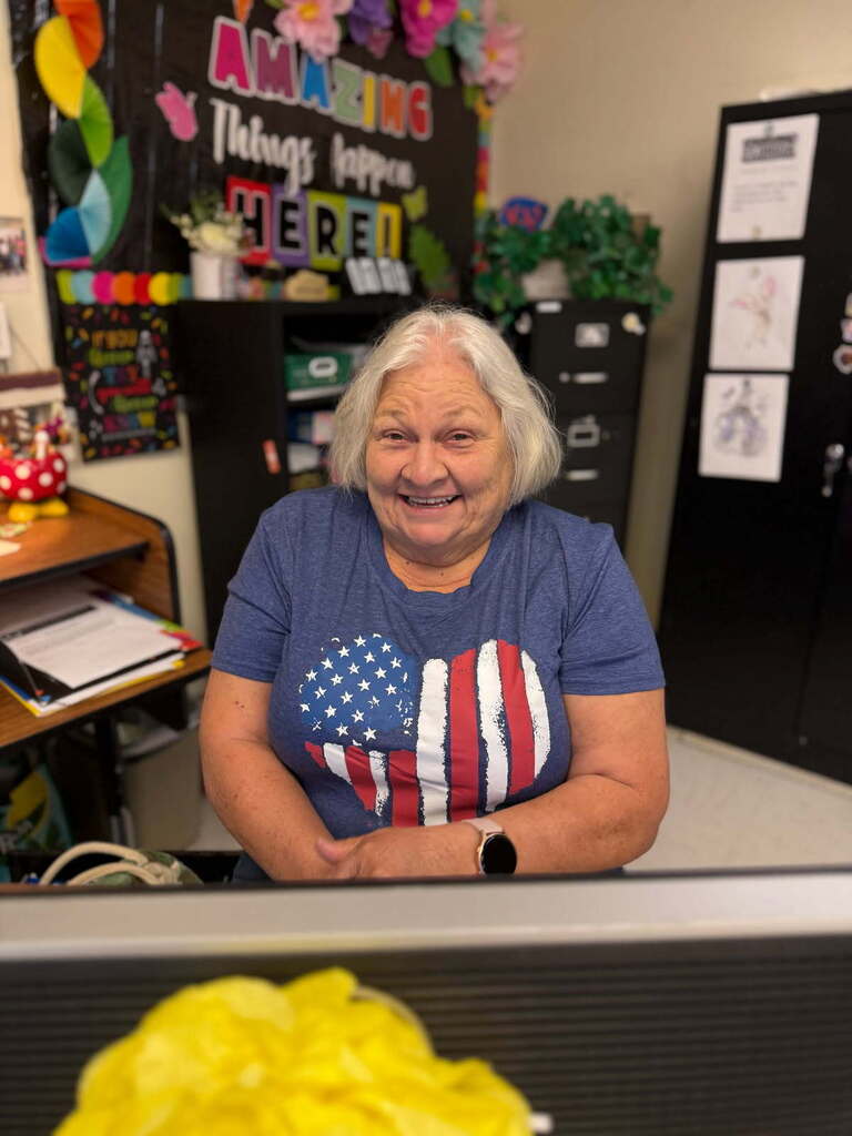 A woman with white hair wearing an American flag heart shirt sits in front of a computer in a classroom.
