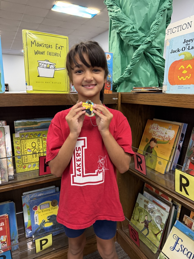 A girl with short hair smiles in a library. She holds a gold coin and wears a red T-shirt. Behind her are shelves with books, one labeled "Fiction."