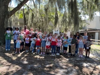 A group of children and adults stand under a tree. They are wearing red shirts and holding certificates.