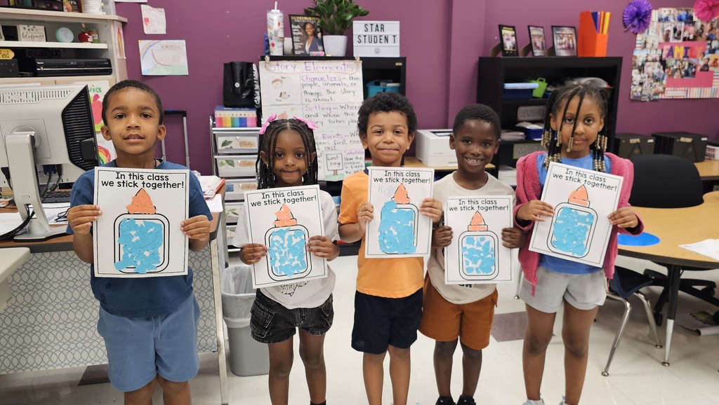 Five children in a classroom hold their cut/glue projects. They are smiling and standing together. 