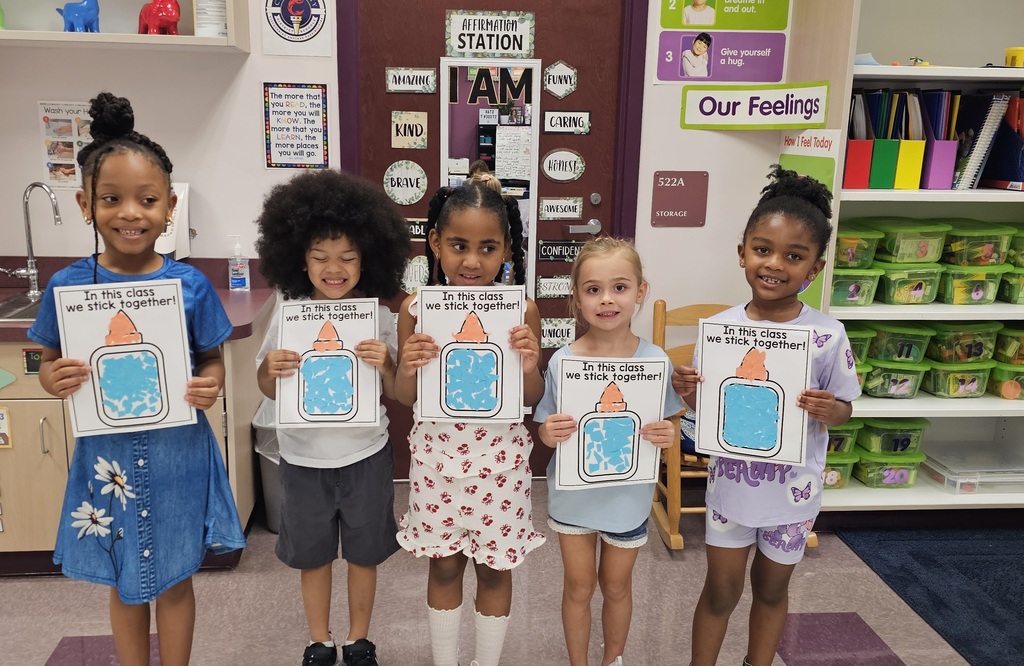 Five children in a classroom hold their cut/glue projects. They are smiling and standing together. 