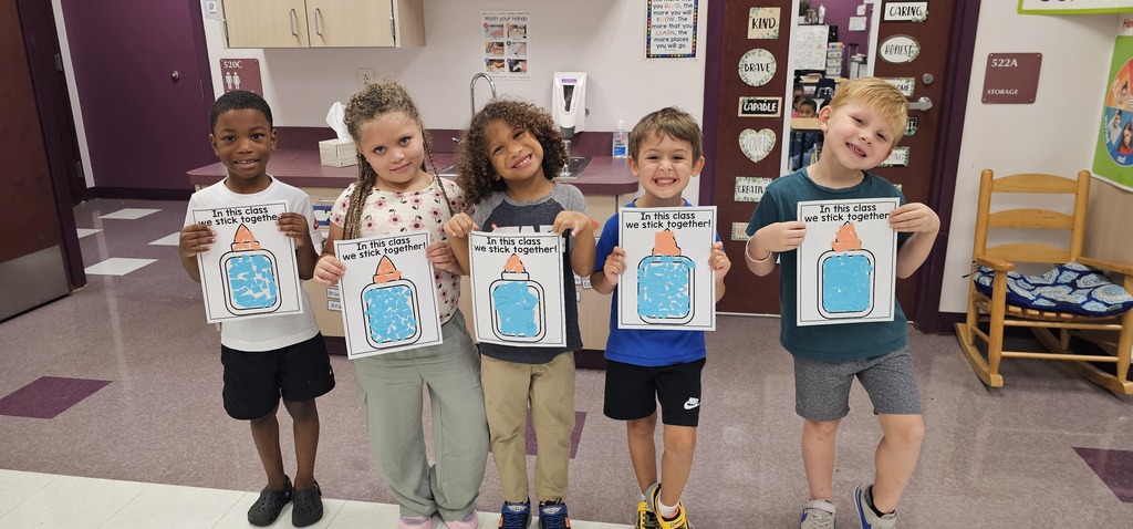 Five children in a classroom hold their cut/glue projects. They are smiling and standing together. 