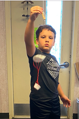 A boy in black shorts and shirt demonstrates a kite while holding a string indoors.