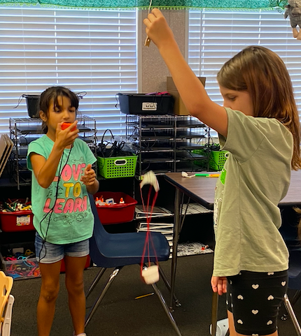 Two young girls are holding strings with a ball, appearing to play indoors in a classroom setting.