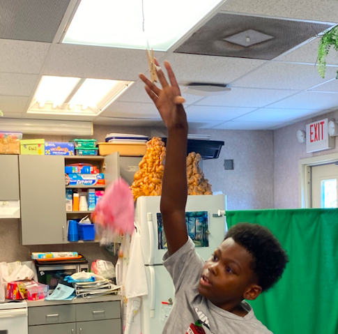 A young boy raising a hand in a classroom with cabinets, a refrigerator, and an exit sign.