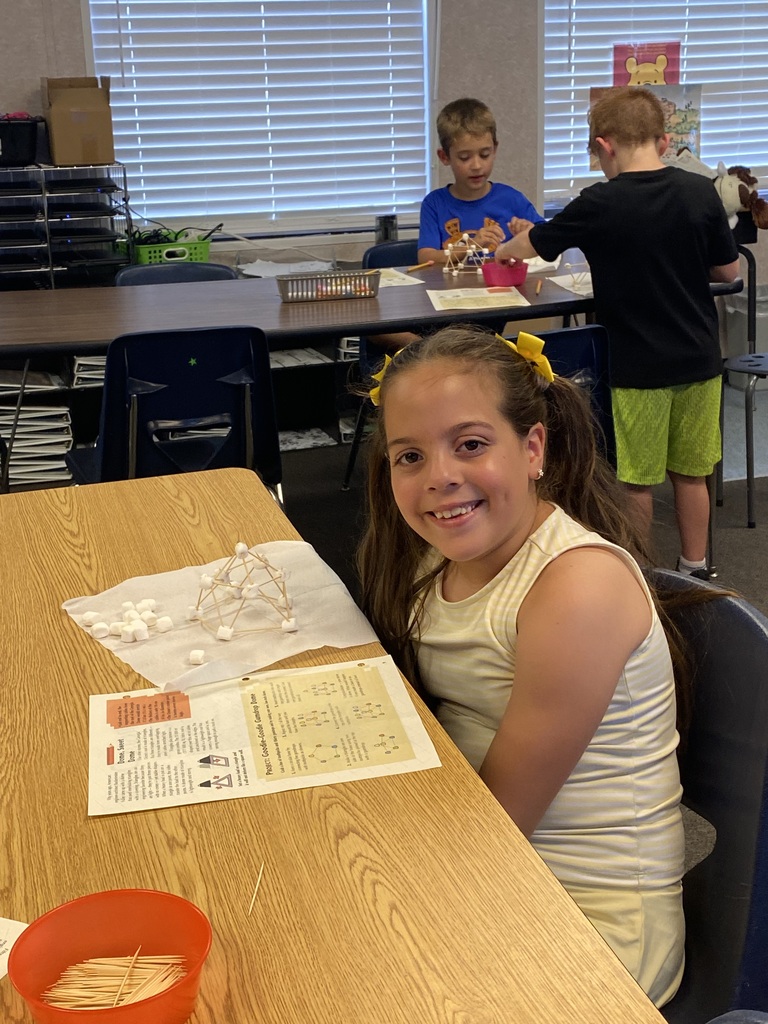 Four kids sit at tables in a classroom. One girl smiles while sitting at a table with a bowl.