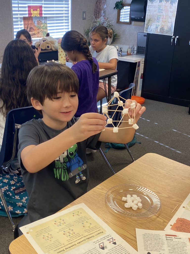 Child holds structure made from marshmallows and toothpicks at a desk. Other children sit at tables.