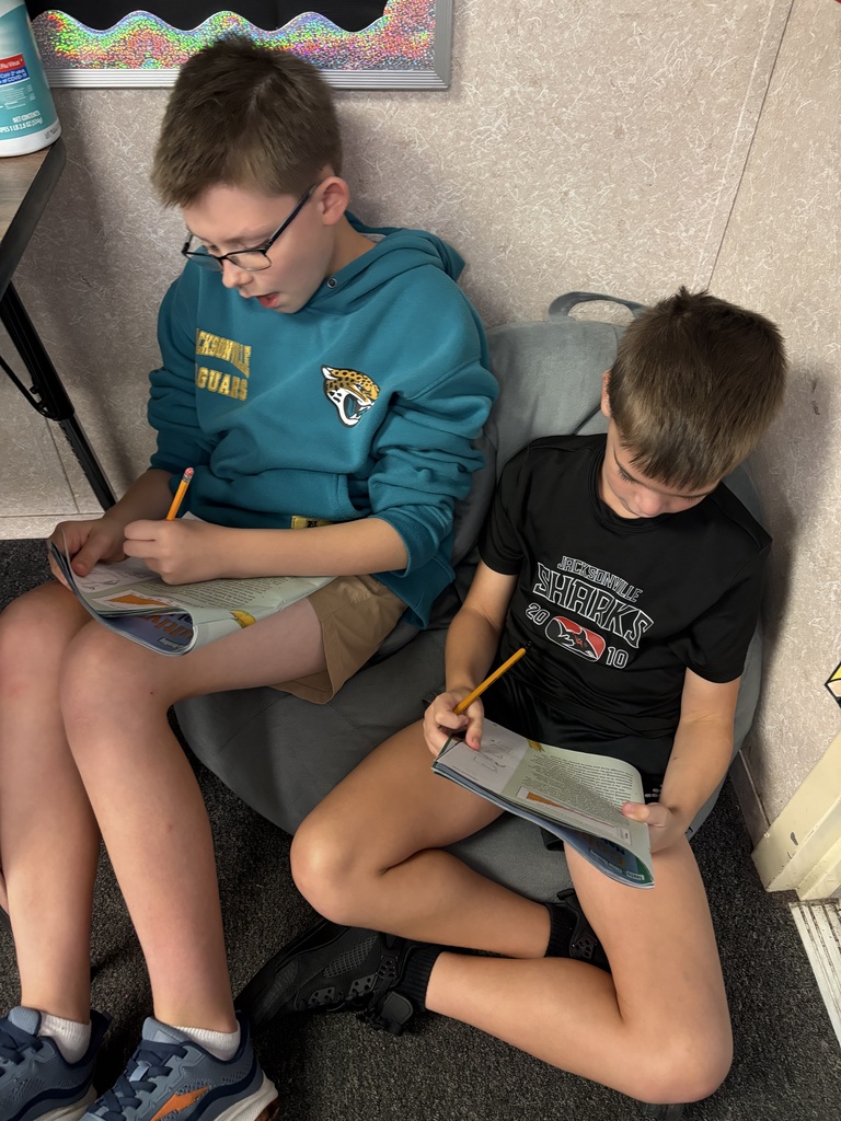 Two boys sitting on bean bags, engaged in reading and writing activities. One holds a book, the other a pencil.