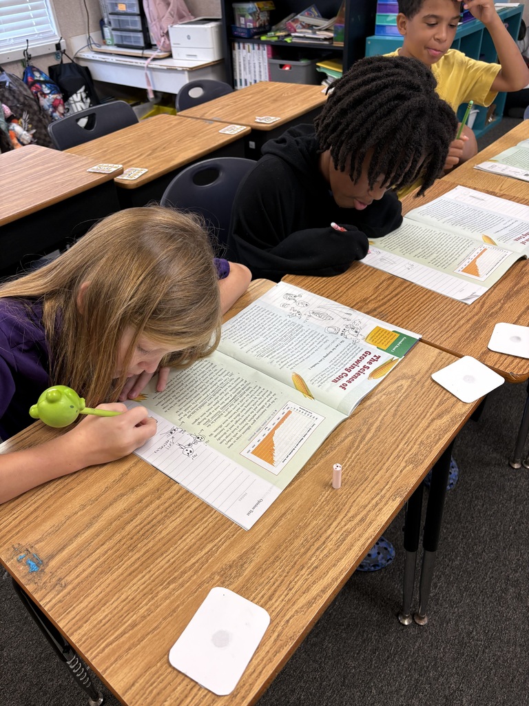 Three children seated at a desk in a classroom, writing in open books. One child has a toy.
