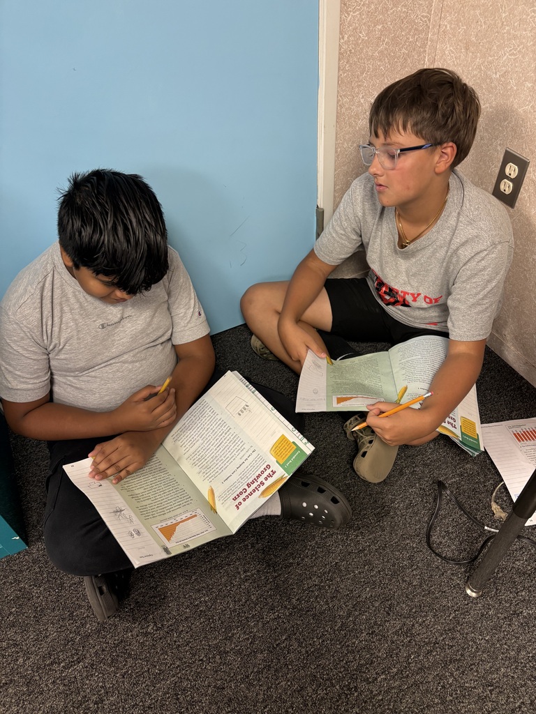 Two students, one with glasses, sit on a floor, reading books and holding pencils.