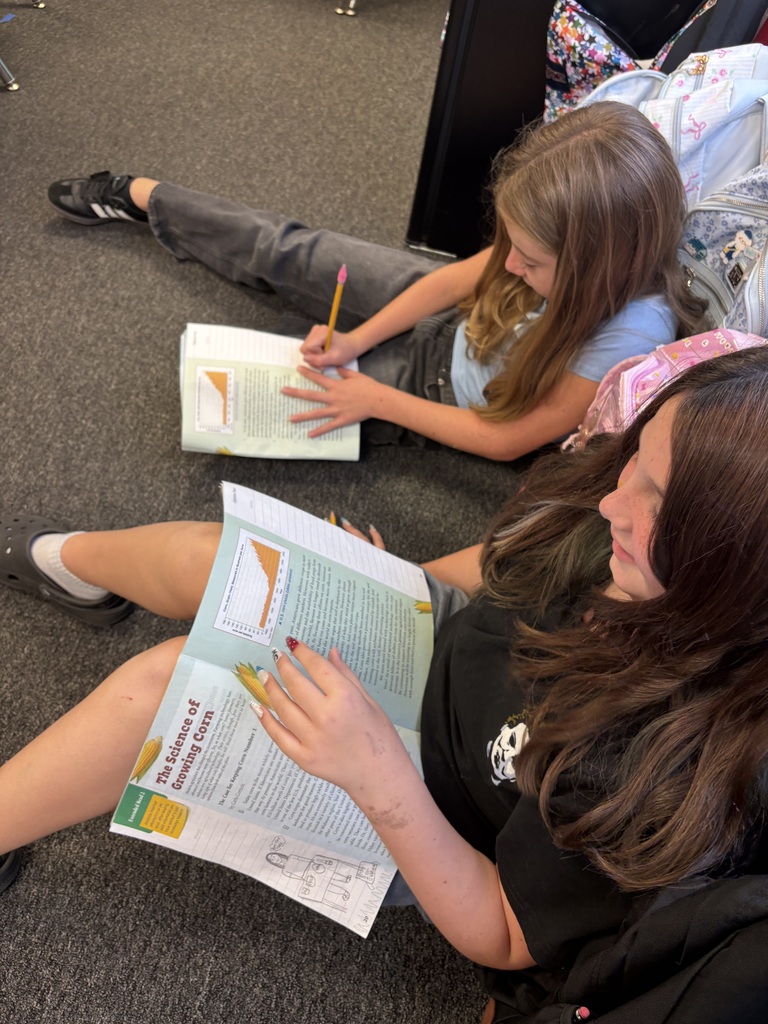 Two students sit on a carpeted floor, one reading a book and the other writing in a book.