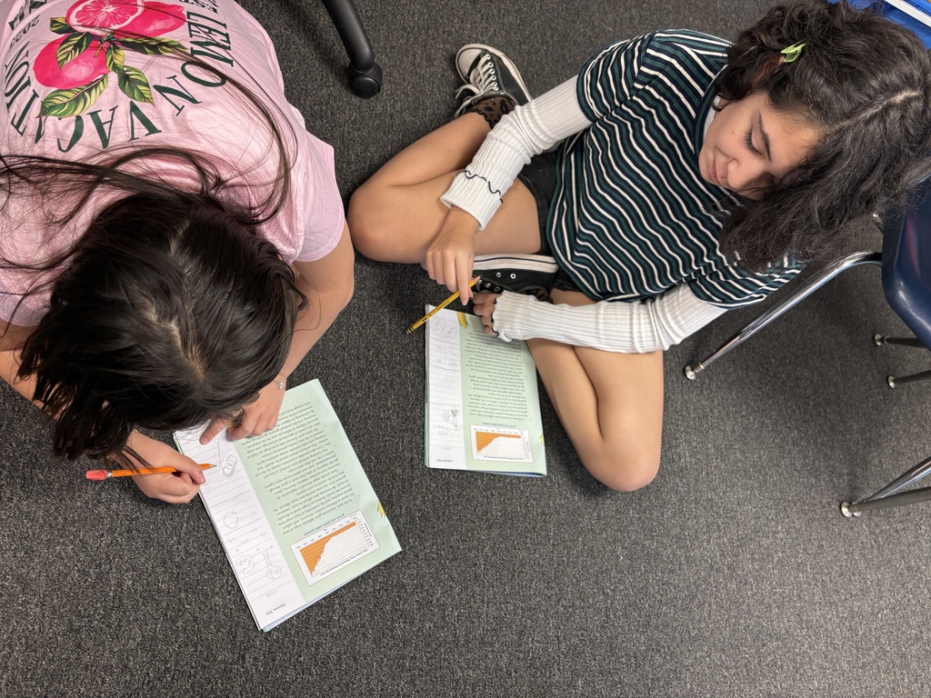 Two girls sitting on the floor with papers and pencils. One girl in pink wears a bracelet.
