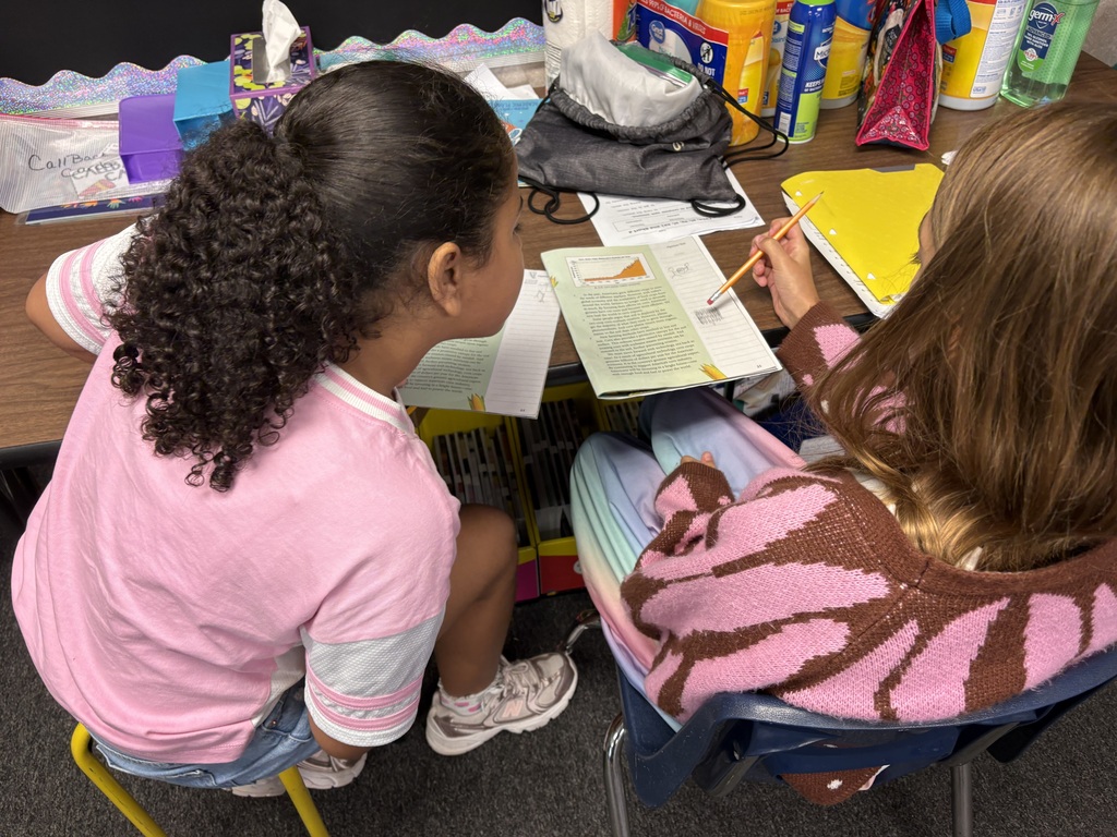 Two young girls in a classroom, one writing and the other reading a book. They sit in chairs.