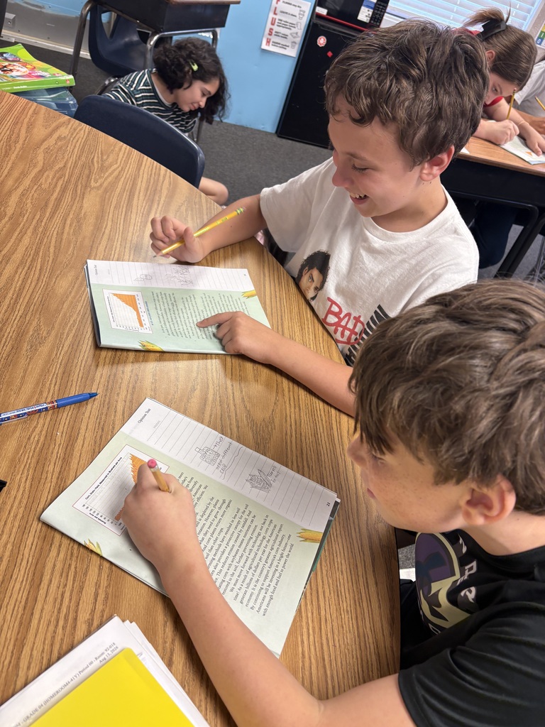 Two students work at a table, one with an open book and pen, the other writing on paper.