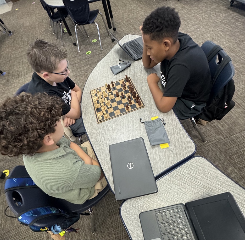 Three young boys seated around a table playing chess, one with curly hair, one with glasses, and another with a backpack.