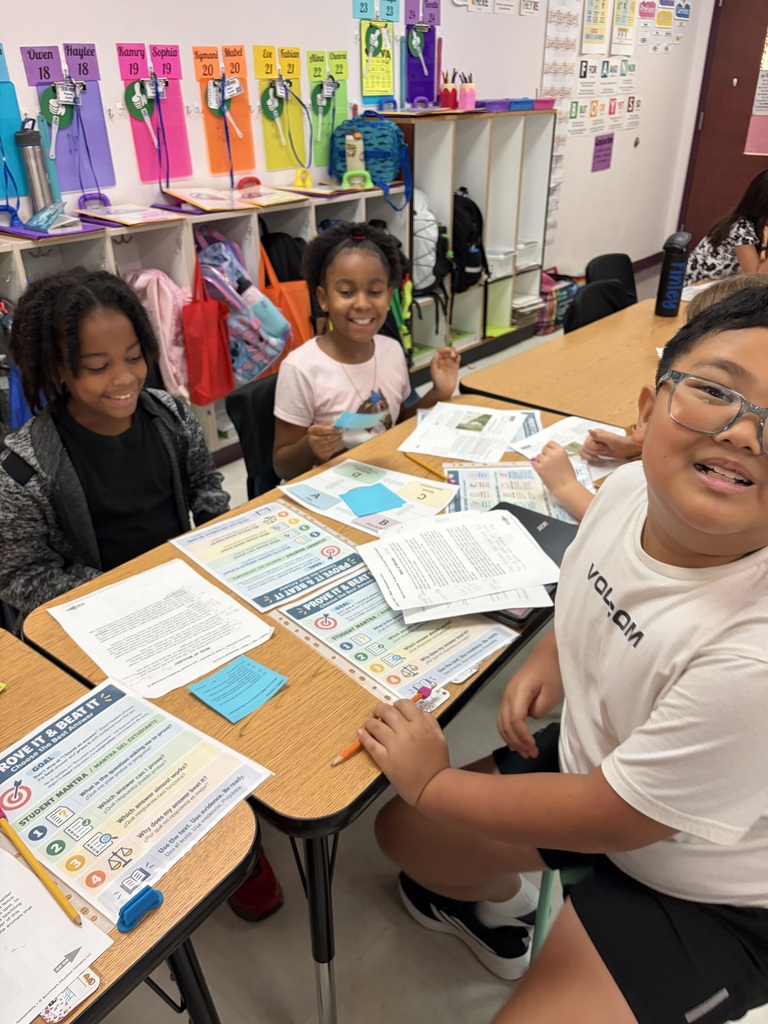 Three children seated at a table in a classroom, surrounded by papers and school supplies.